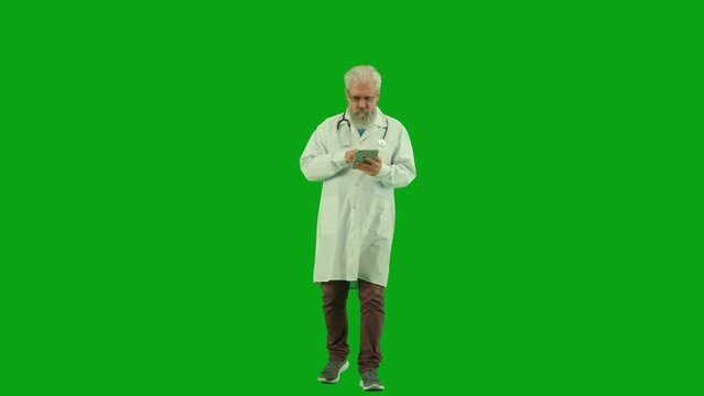 Portrait of aged man medic on chroma key green screen. Senior doctor in uniform walking and holding tablet, texting looking information online.