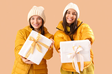 Young women in winter clothes with Christmas gifts on beige background