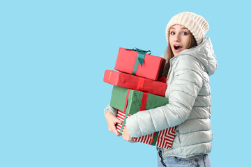 Surprised young woman in winter clothes with Christmas gifts on blue background