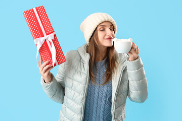 Young woman in winter clothes with cocoa and Christmas gift on blue background