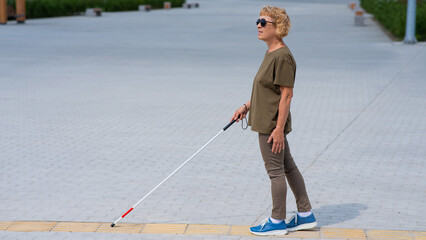 An elderly blind woman walks with a cane along a tactile tile. 