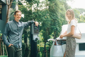 Young couple travel with EV electric car charging in green sustainable city outdoor garden in...