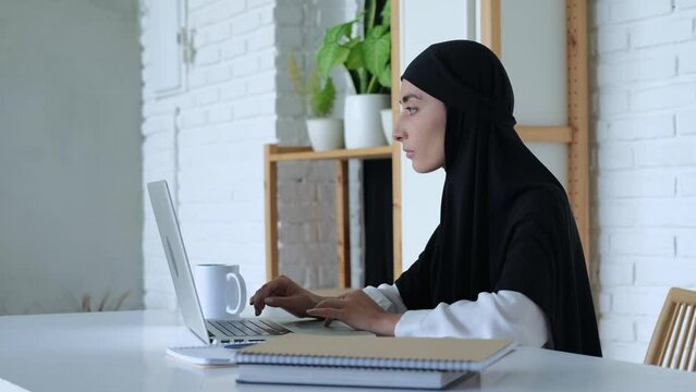 Muslim student wearing hijab is typing dissertation on computer. Student is working intently on task. Working on laptop makes it easier for students to create new printed publications.