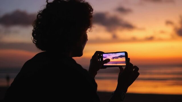 Woman captures brightly colored sky with her phone camera and magnificent sunset by sea. Screen reflects soft lighting and rich shades of sky. Sky and sunset become source of inspiration for person.