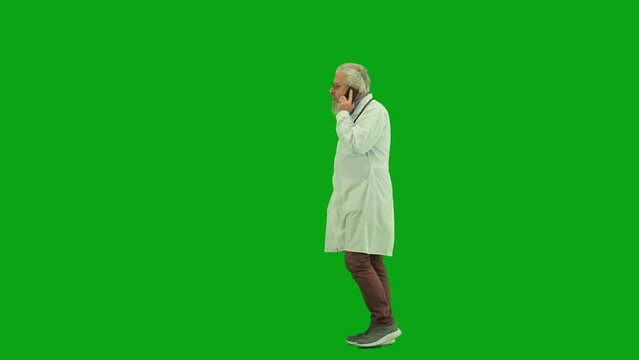 Portrait of aged man medic on chroma key green screen. Side view senior doctor walking through clinic or hospital and talking on smartphone.