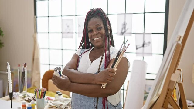 Confident african american woman artist with braids, paintbrushes in hand, standing with crossed arms gesture inside beautiful art studio