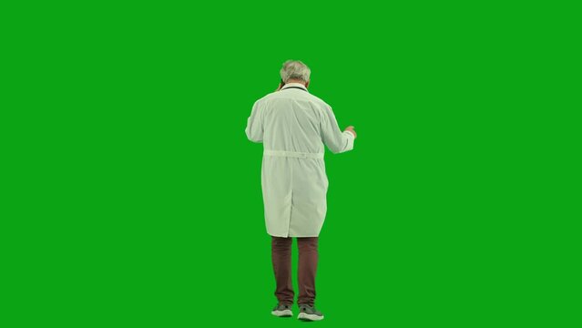 Portrait of aged man medic on chroma key green screen. Back view senior doctor in uniform walking in hospital corridor talking on smartphone.
