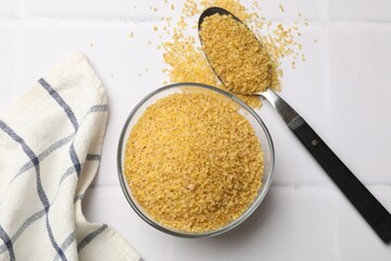 Bowl and spoon with raw bulgur on white tiled table, top view