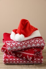Stack of different Christmas sweaters and Santa hat on wooden table against beige background