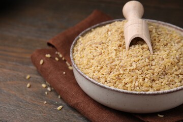 Bowl and scoop with raw bulgur on wooden table, closeup. Space for text