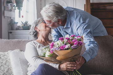 Old man giving flowers at his wife sitting on the sofa at home for the San Valentines’ day. Pensioners enjoying surprise together. In love people having fun..
