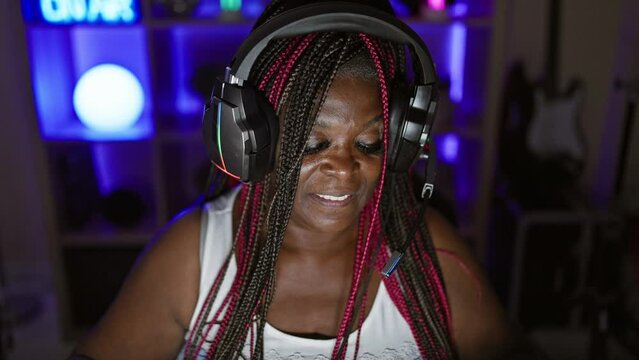 Glowing african american woman streamer in the heart of the gaming room, rocking a night session, stretching arms amidst digital entertainment, beautiful portrait of a gamer queen wearing headphones
