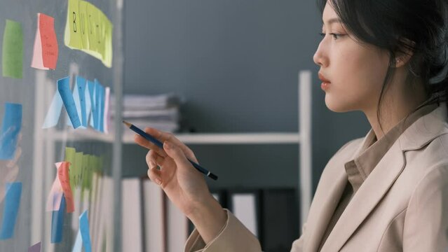 Young smiley attractive businesswoman using sticky notes in glass wall to writing strategy business plan to development grow to success