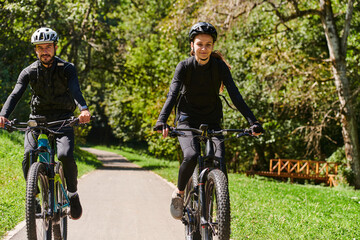 Naklejka premium A blissful couple, adorned in professional cycling gear, enjoys a romantic bicycle ride through a park, surrounded by modern natural attractions, radiating love and happiness