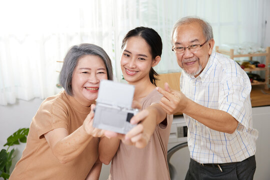 asian family senior couple with granddaughter talking selfie from digital camera, making miniheart sign and duck face at home