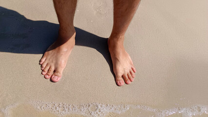 Top view of bare men's feet with approaching seawave on the shore with selective focus, Low angle...