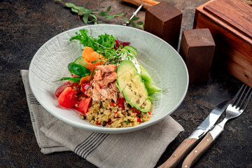 Salad with canned tuna, avocado, tomatoes and bulgur in a white plate. Served on a brown background