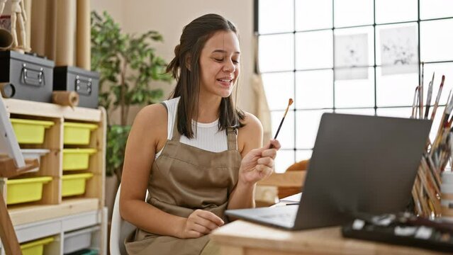 Cheerful young hispanic woman artist beaming during an inspiring online art class while enjoying a video call at her cozy studio