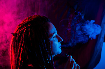 Profile of caucasian girl with dreadlocks smokes a vape in red blue light. 