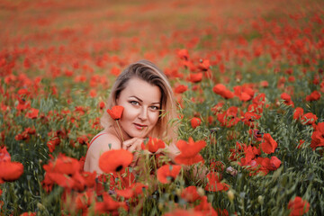 Naklejka premium Happy woman in a red dress in a beautiful large poppy field. Blond sits in a red dress, posing on a large field of red poppies