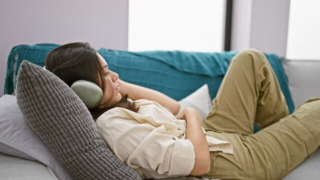 Young beautiful hispanic woman using smartphone and headphones lying on sofa at home