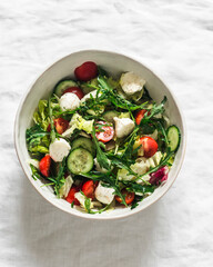 Arugula, cherry tomatoes, cucumbers, mozzarella cheese salad with lemon-mustard dressing on a light background, top view