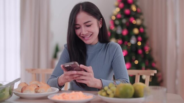 Asian young women are using their mobile phones at the dining table in the festive atmosphere of Christmas celebration at her home. Find the perfect stock image to capture the holiday spirit.