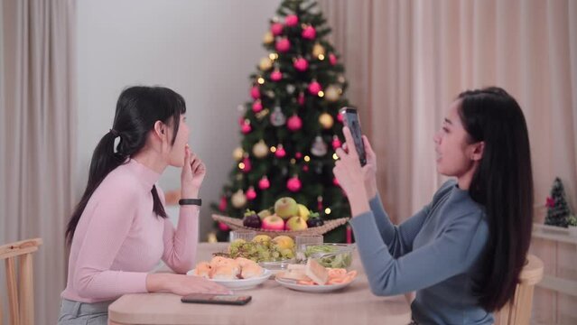 Asian female friends capture memories by taking photos together using smartphones at the dining table in the festive atmosphere of Christmas at her home.