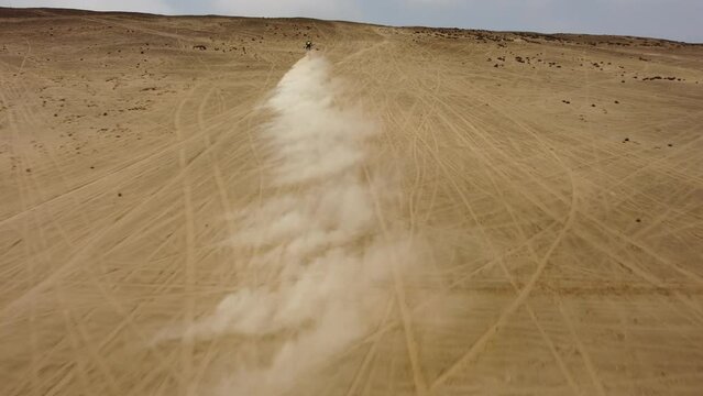 Motorcyclist quickly riding up a steep desert hill blowing sand in the air. Drone follows behind and flies upwards revealing more dunes and mountains in the background horizon. Located in Lima, Peru.