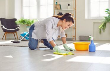 Happy housewife cleaning the floor at home. Young woman in rubber gloves washing and wiping light gray wooden or laminate flooring in the living room. Housework concept