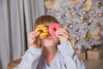 cheerful young woman holds donuts in her hands on christmas tree background, winter holidays. portrait of funny girl in pajamas covering her eyes with donuts