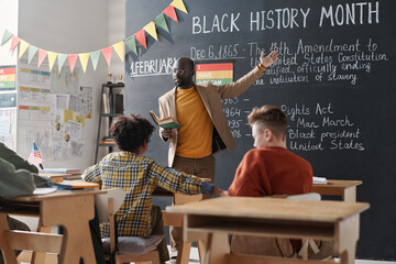African AMerican teacher pointing at blackboard and talking about Black History Month to school...