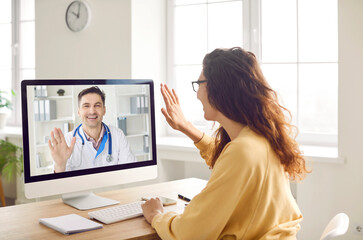 Patient says hello to online doctor when they start medical consultation via video call. Woman sitting in front of computer screen and waving her hand to greet her remote doctor. Telemedicine concept - obrazy, fototapety, plakaty