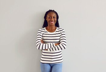Portrait of beautiful female model in casual clothes. Happy young African American woman wearing white black striped long sleeve shirt and blue jeans standing with folded arms by light gray wall - Powered by Adobe