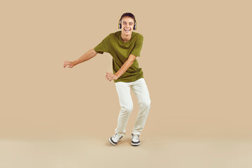 Fototapeta na wymiar Full length portrait of a young joyful boy wearing casual clothes having fun, dancing and listening to music in headphones, looking at camera isolated on a studio beige background.