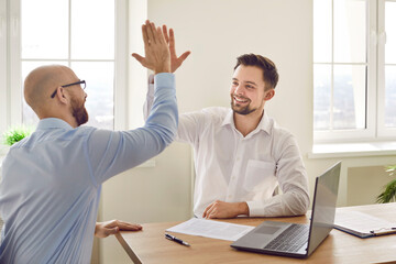 Two happy smiling young colleagues business men giving high five at meeting sitting at the desk on...