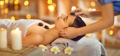 Close up portrait of a pretty young brunette woman with closed eyes lying relaxing in spa salon getting face and head massage therapy with hot stones. Wellness and beauty day concept. Banner.