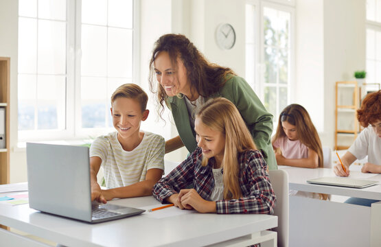 Happy school children using a laptop in the classroom. Cheerful schoolboy and schoolgirl together with their friendly teacher looking at the notebook computer and smiling. Technology education concept