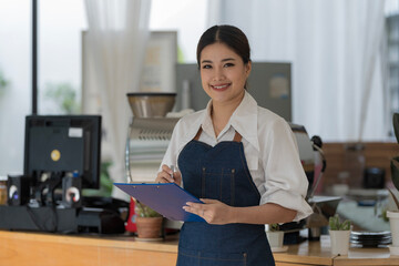 A young Asian barista holding a menu folder stands in front of a coffee shop counter, looks at the...