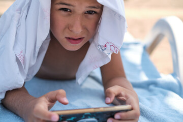 A boy lounges absorbed in his phone, with a sandy beach backdrop. Emphasizes the digital invasion...