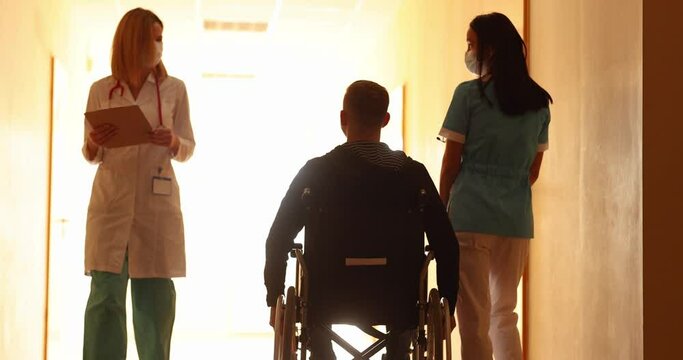 Nurse walking with disabled man in wheelchair along corridor of clinic and greeting doctor 4k movie. Rehabilitation of people with disabilities concept