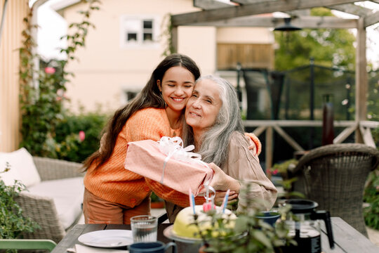 Happy girl embracing and giving birthday gift to grandmother at patio