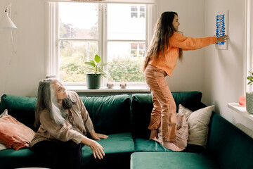 Girl arranging painting on wall while grandmother sitting in sofa at home