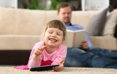 Little smiling baby girl with smartphone lies on floor in background dad reads a book. Children...