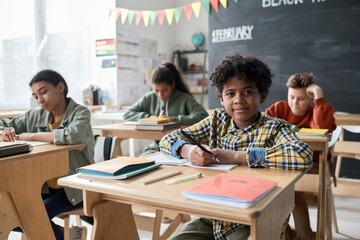 Portrait of schoolboy sitting at desk in the classroom and smiling at camera with his classmates in the background
