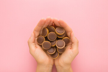 Brown caramel chocolate candies in young adult woman opened palms on light pink table background....