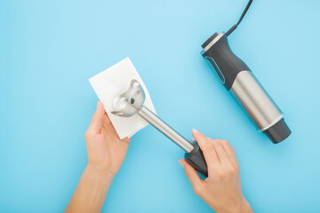 Young adult woman hands cleaning immersion blender with dry white paper napkin on light blue table background. Pastel color. Closeup. Home kitchen appliance purity. Point of view shot. Top down view.