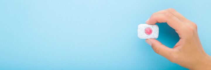 Young adult woman hand holding and showing blue red dishwasher tablet on light blue table background. Pastel color. Closeup. Wide banner. Empty place for text. Top down view.