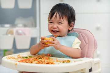 happy infant baby eating food and vegetable by self feeding BLW or baby led weaning on chair