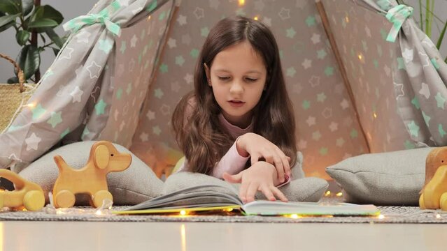 Curious little girl with dark hair reading paper book turning pages looking at pictures sitting on floor in wigwam at home enjoying new textbook with stories.